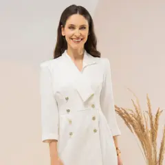 Uśmiechnięta kobieta w eleganckiej, białej sukience tuba z dwurzędowym zapięciem i złotymi guzikami.