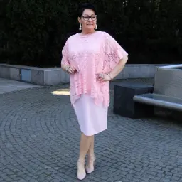 Elegancka kobieta w różowej sukience plus size z koronkową narzutką, stojąca na brukowanym placu.