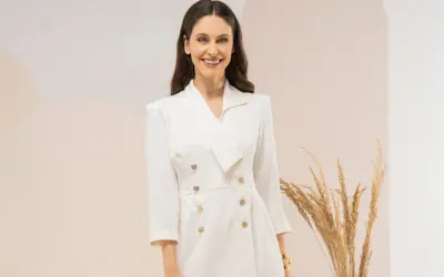 Uśmiechnięta kobieta w eleganckiej, białej sukience tuba z dwurzędowym zapięciem i złotymi guzikami.
