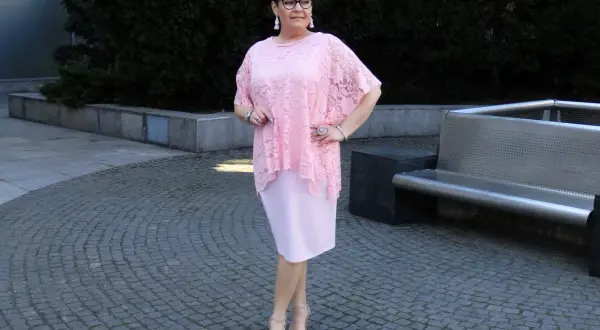 Elegancka kobieta w różowej sukience plus size z koronkową narzutką, stojąca na brukowanym placu.