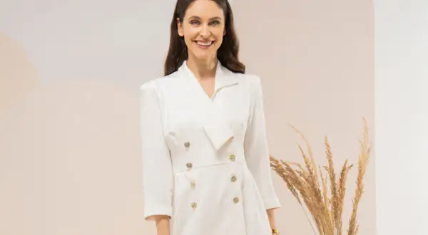 Uśmiechnięta kobieta w eleganckiej, białej sukience tuba z dwurzędowym zapięciem i złotymi guzikami.
