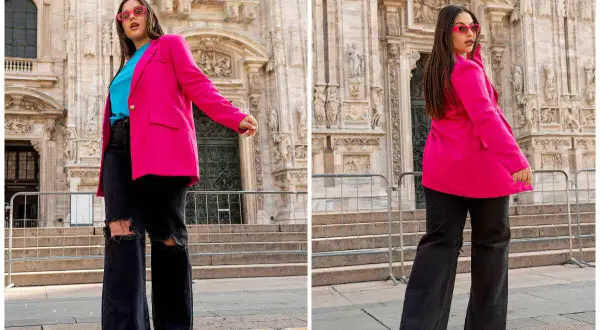Kobieta w różowej, długiej marynarce damskiej w dużym rozmiarze, czarnych jeansach z rozdarciami i turkusowym topie. Stylowy look na tle zabytkowej architektury.