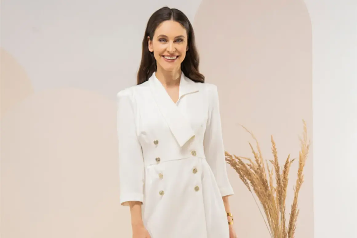 Uśmiechnięta kobieta w eleganckiej, białej sukience tuba z dwurzędowym zapięciem i złotymi guzikami.